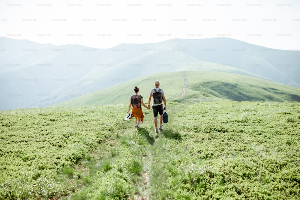 Coppia che cammina con gli zaini sul prato verde, viaggiando in montagna durante il periodo estivo, ampia vista sul paesaggio