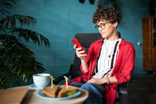Young brunette female with smartphone surfing in social networks while relaxing in cafe by served table
