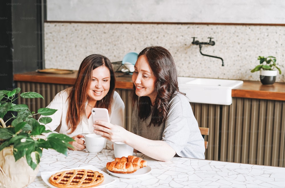 Erwachsene lächelnde brünette Freundinnen frühstücken und benutzen Handy in der Küche im Haus