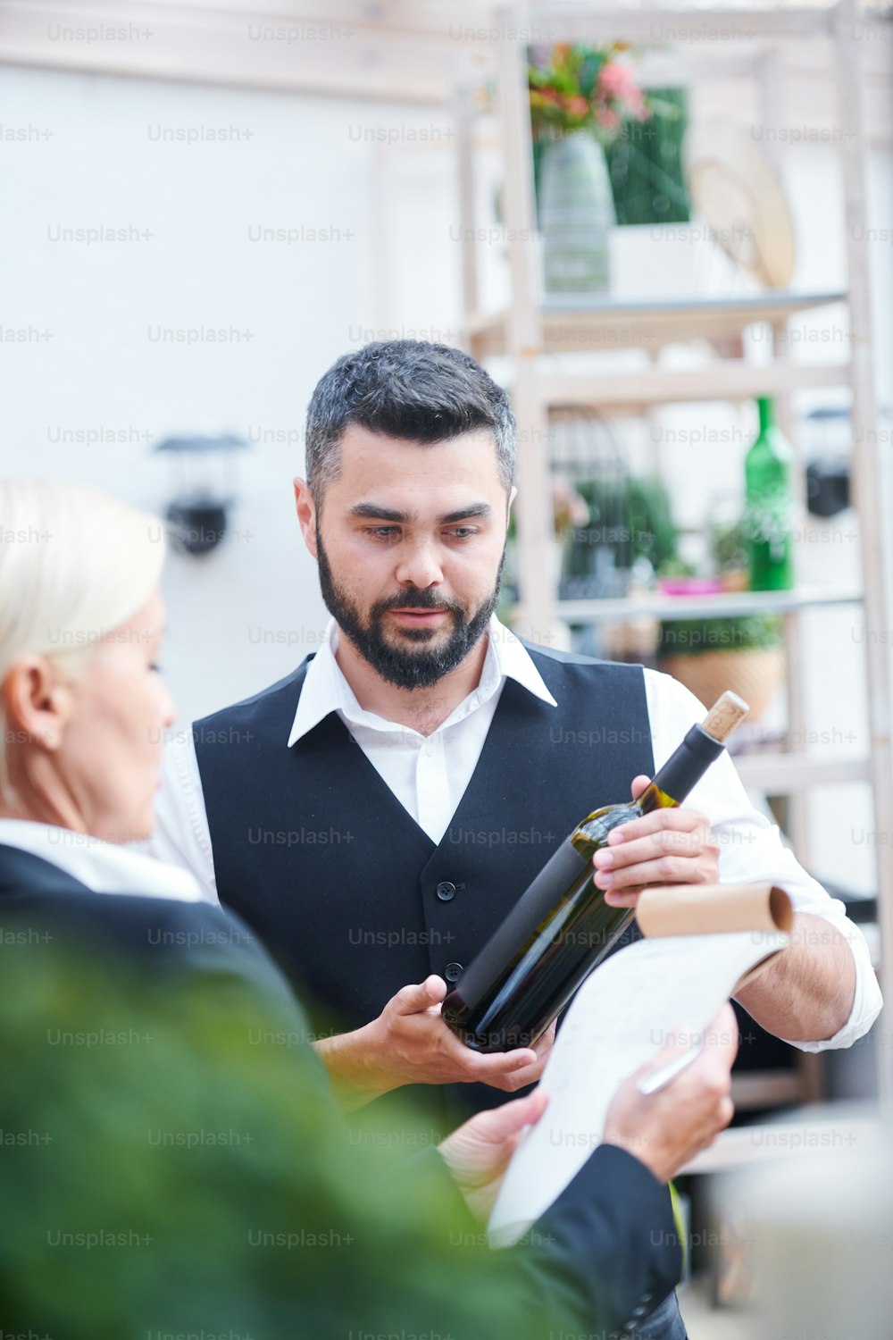 Young bearded bartender giving characteristics of new sort of wine in bottle to cavist while both working in cellar