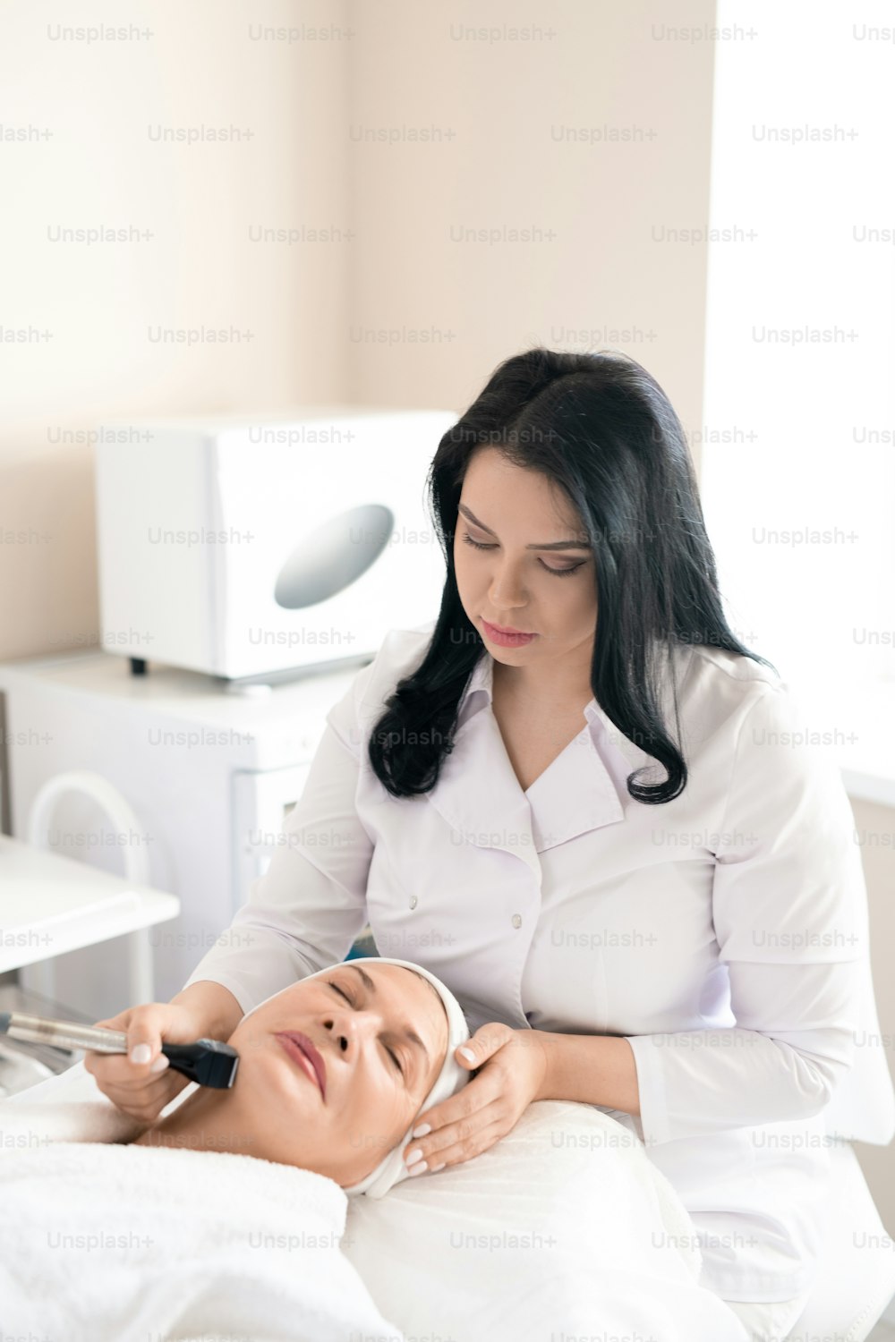 Concentrated cosmetologist in white coat using facial machine while doing skin tightening procedure to mature woman