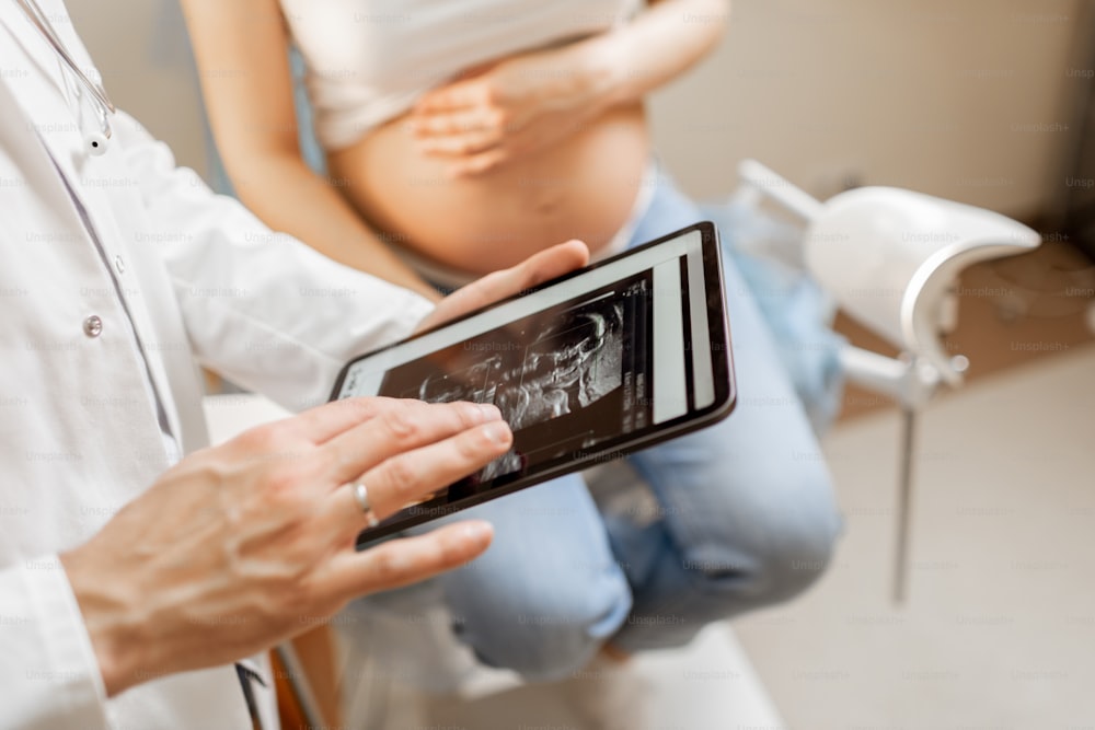 doctor with an ultrasound scan of unborn child on a digital tablet during an examination with a pregnant woman in the office, cropped view without faces
