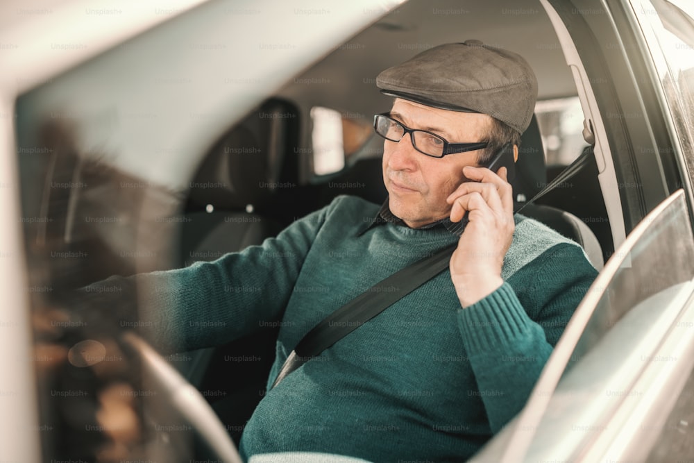 Kaukasischer Senior mit Hut auf dem Kopf und Brille fährt Auto und benutzt Smartphone.