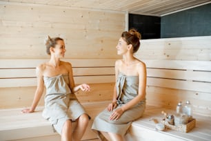 Two young girlfriends sitting and talking together while relaxing in the sauna. Concept of female friendship and spa treatment