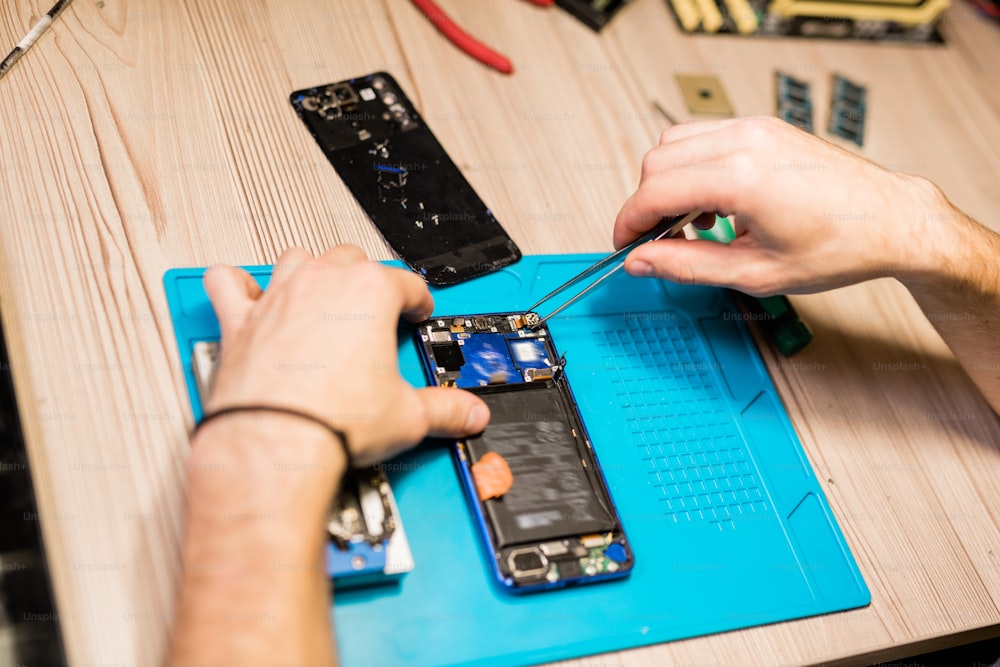 Hände des Reparateurs über kaputtes Gerät mit Pinzette, um winzige Teile oder Schrauben des demontierten Smartphones zu reparieren