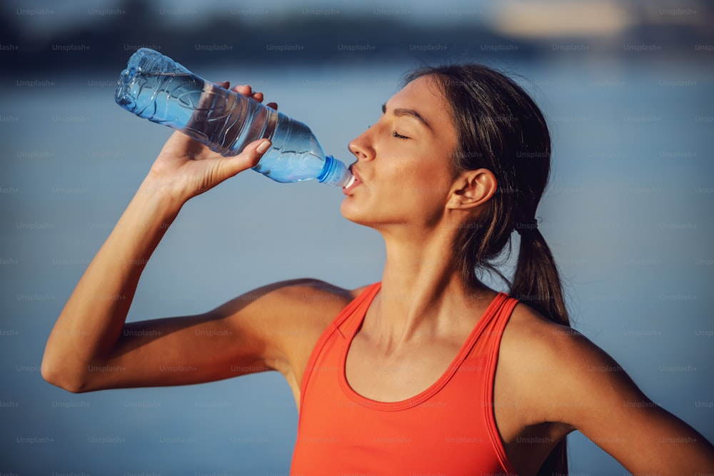 Profil einer attraktiven, schlanken, durstigen Sportlerin, die frisches Wasser aus der Flasche trinkt.