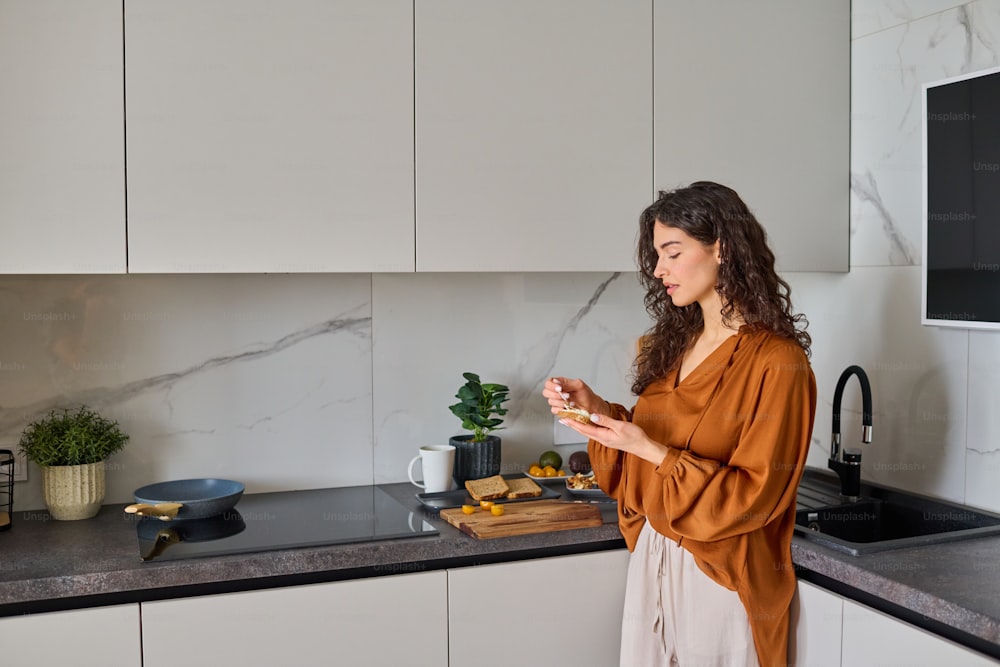 Young healthy female in blouse and pants spreading cheese on slice of bread while making sandwich for breakfast in the kitchen