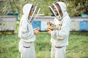 Dos apicultores con uniforme protector de pie junto con miel en el frasco, degustando productos frescos en el colmenar al aire libre