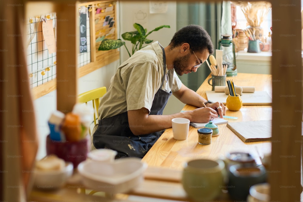Junger kreativer Mann, der sich über den Arbeitsplatz beugt, während er im Notizblock in einem geräumigen Studio oder einer Werkstatt mit Displays für neue Gegenstände skizziert
