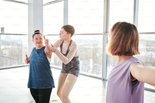 Giovane istruttore di yoga e fitness in abbigliamento sportivo in piedi accanto a una ragazza disabile e aiutandola con uno degli esercizi durante l'allenamento fisico in palestra