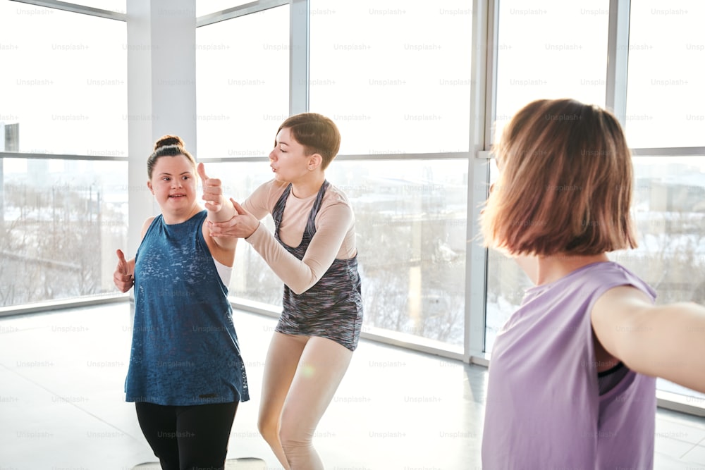 Young yoga and fitness trainer in activewear standing by disable girl and helping her with one of exercises during physical training in gym