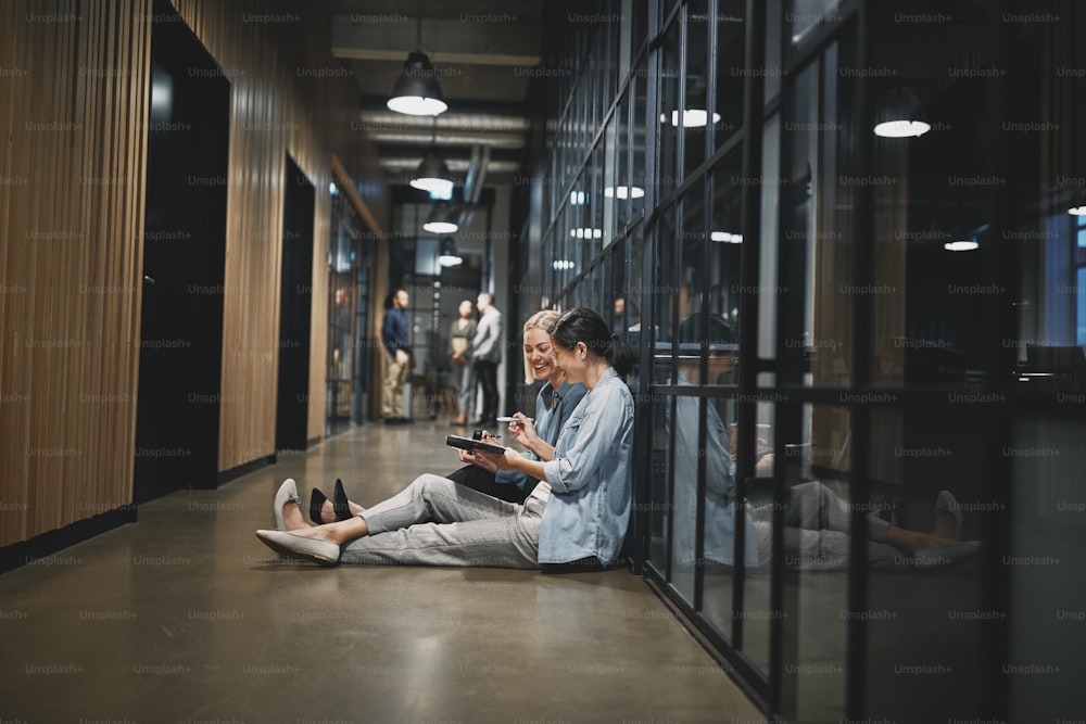 Two smiling young businesswomen talking while sitting together in the hallway of an after a late meeting