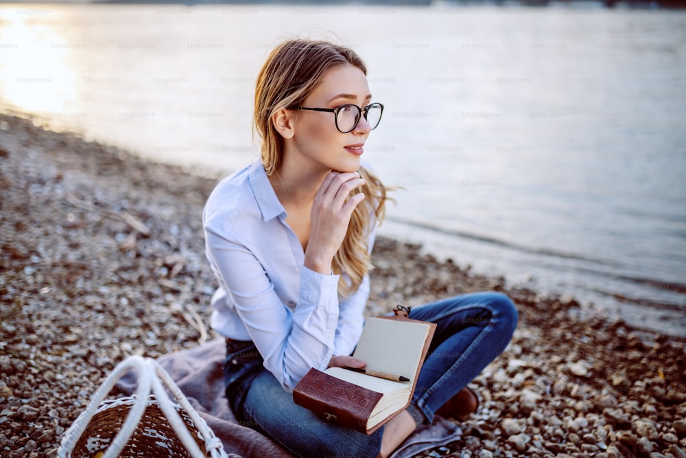 Attractive cute caucasian fashionable young woman with eyeglasses sitting on coast near river, thinking and holding notebook.