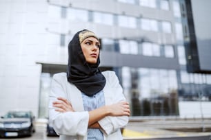Attraktive, selbstbewusste muslimische Geschäftsfrau steht mit verschränkten Armen vor ihrer Firma.