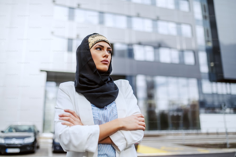 Empresária muçulmana atraente e confiante de pé na frente de sua empresa com os braços cruzados.