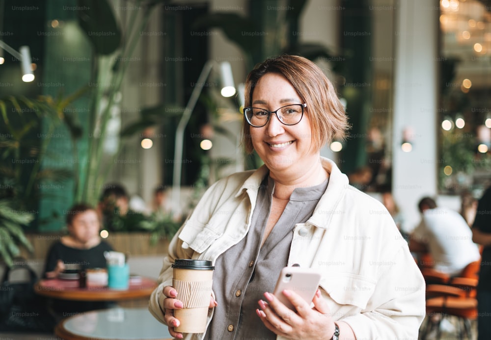 Adult brunette middle aged woman plus size body positive in casual clothes with paper cup of coffee using mobile phone in cafe