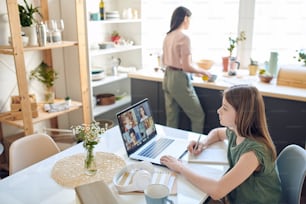 Concentrated teenage daughter sitting at desk and using laptop while participating in group video conferencing with tutor