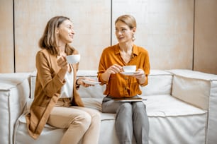 Two female colleagues having cheerful conversation, sitting together on the couch during a coffee break in the office