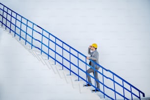 Full length of handsome caucasian businessman with helmet on head climbing oil storage tank.