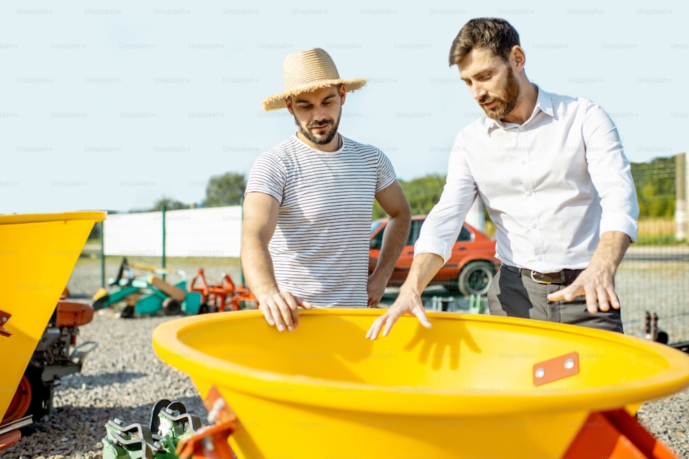 Young agronomist with salesman at the open ground of the shop with agricultural machinery, buying a new planter for farming