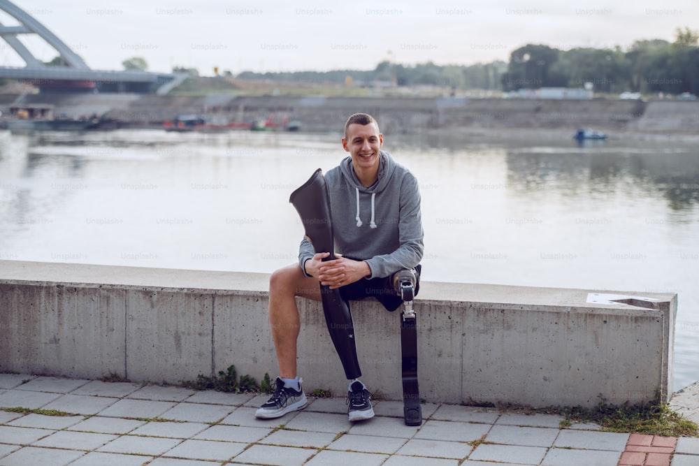 Handsome sporty caucasian handicapped man in sportswear and with artificial leg sitting on quay and holding another artificial leg.