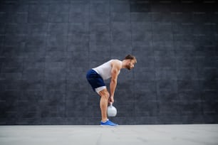 Strong muscular handsome Caucasian man in shorts and t-shirt standing outdoors and lifting kettle bell. In background is gray wall.