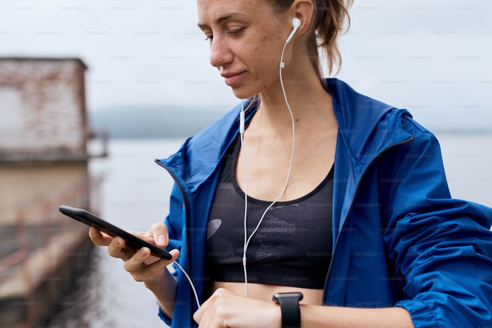 Portrait of mid age skinny sports woman with earphones synchronizing smart watch with smart phone outdoor in summer, on gloomy day with scenic view, wearing tank and blue rain coat