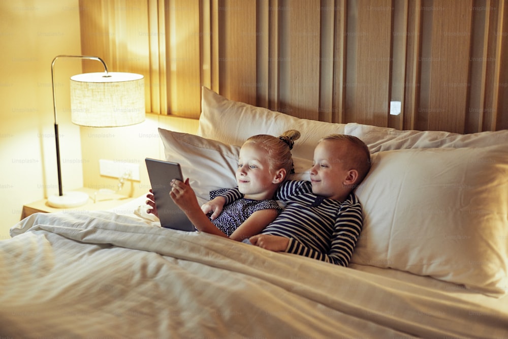 Cute little brother and sister lying in bed together watching a video on a digital tablet before bedtime