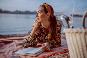 Attractive caucasian brunette in floral dress and with headband lying on stomach on blanket on the shore and reading book. In foreground is picnic basket.