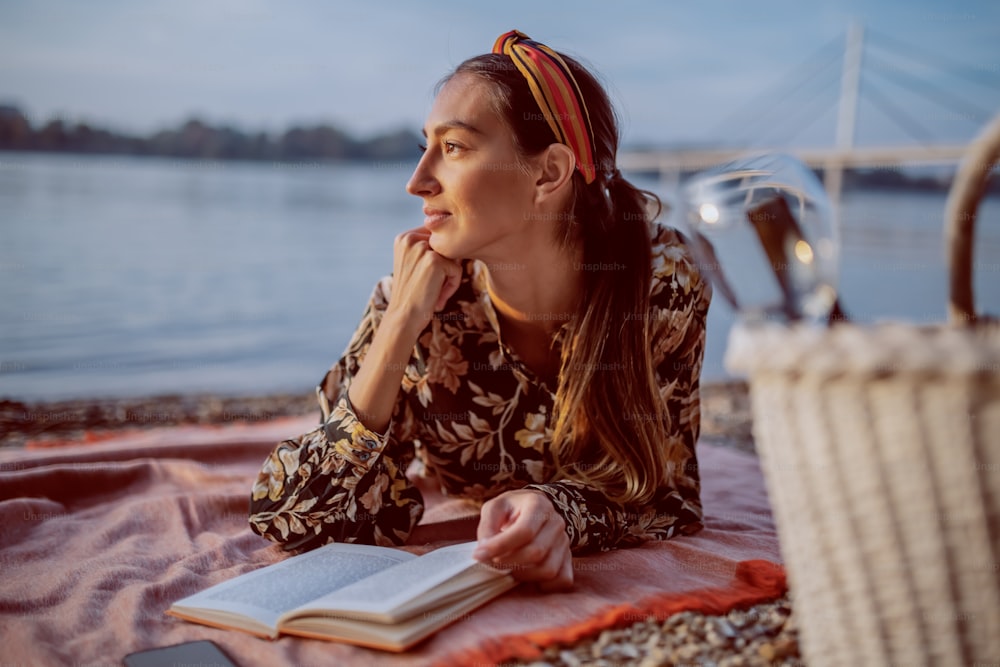 Atractiva morena caucásica en vestido floral y con diadema acostada boca abajo sobre una manta en la orilla y leyendo un libro. En primer plano está la canasta de picnic.