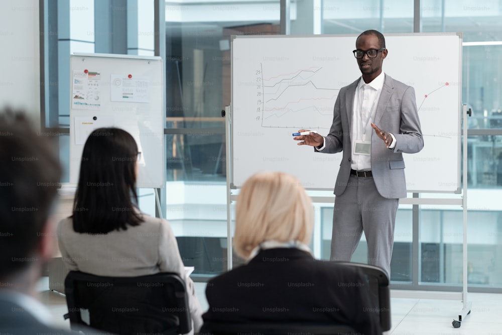 Professor confiante em terno cinza elegante explicando dados gráficos do público no quadro branco durante a palestra de economia na sala de conferências