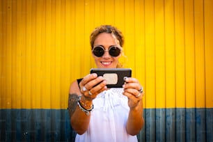 Cheerful adult beautiful woman taking selfie picture with modern phone and urban yellow and blue background - modern people enjoying social media technology  and internet connection outdoor