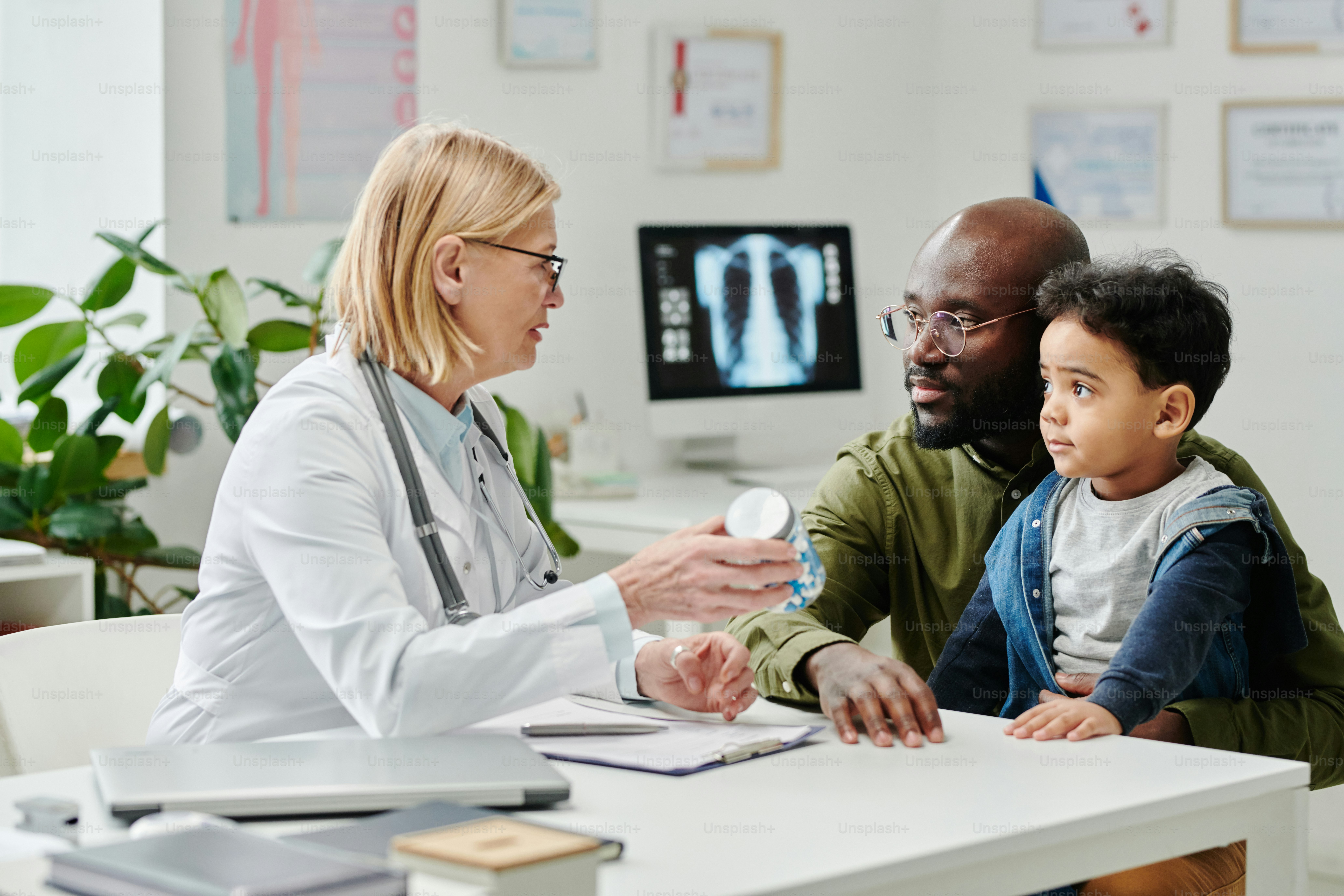 Confident experienced doctor prescribing new medicament to young man with his little son sitting by desk in front of her in clinics