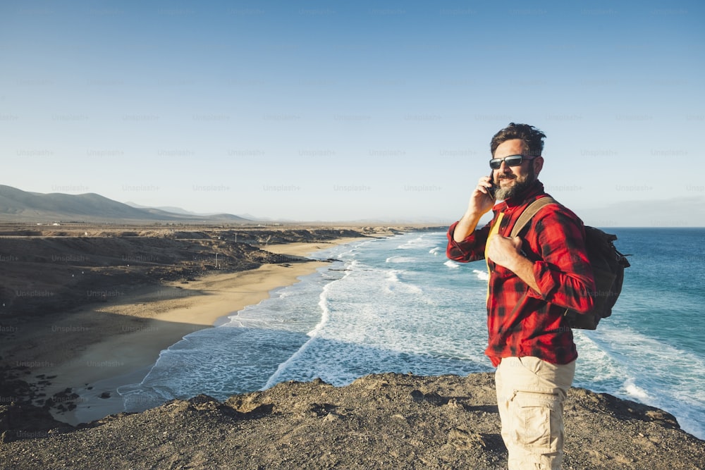 Standing caucasian man calling at the phone on a isolate cliff with ocean waves view - backpacker alternative lifestyle vacation for people love nature and outdoors - technology and adventure concept