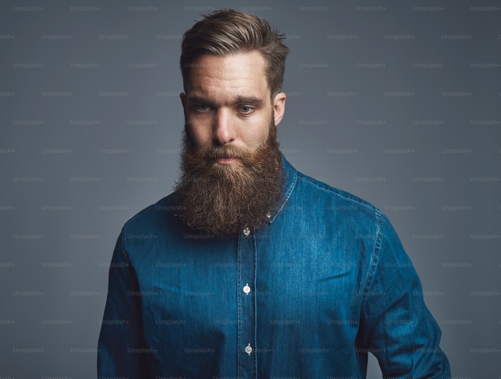 Hip and stylish young man with a long beard looking deep in thought while standing alone against a gray background