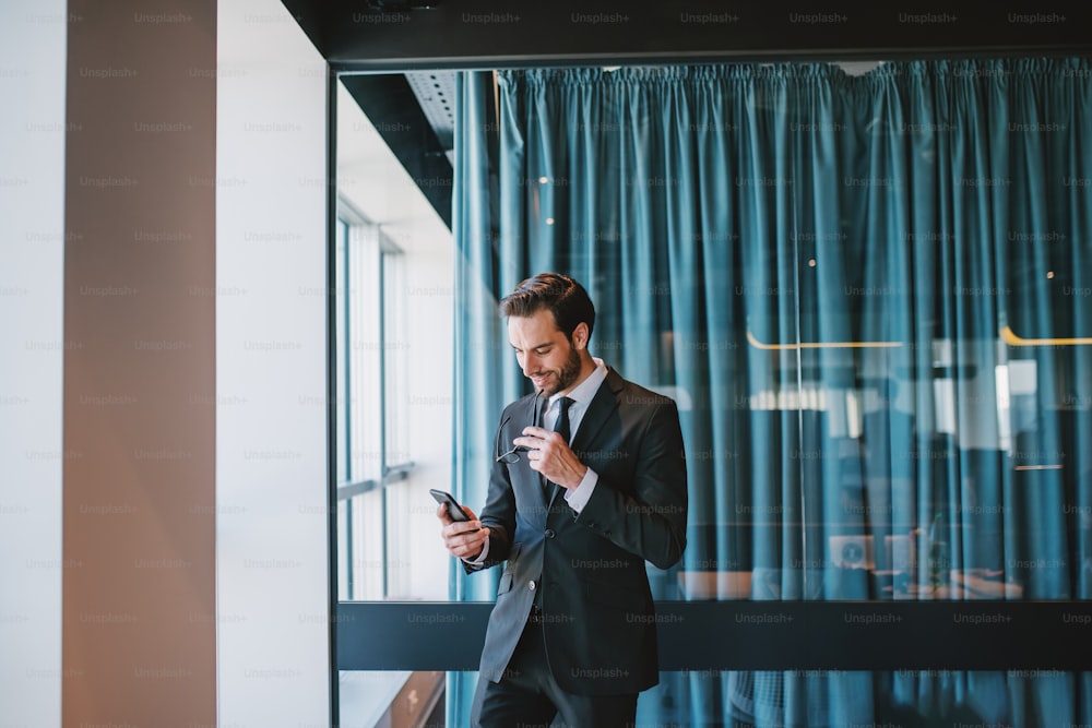 Three quarter length of attractive elegant caucasian businessman in suit holding eyeglasses and reading message on smart phone while standing next to window. Corporate company interior.