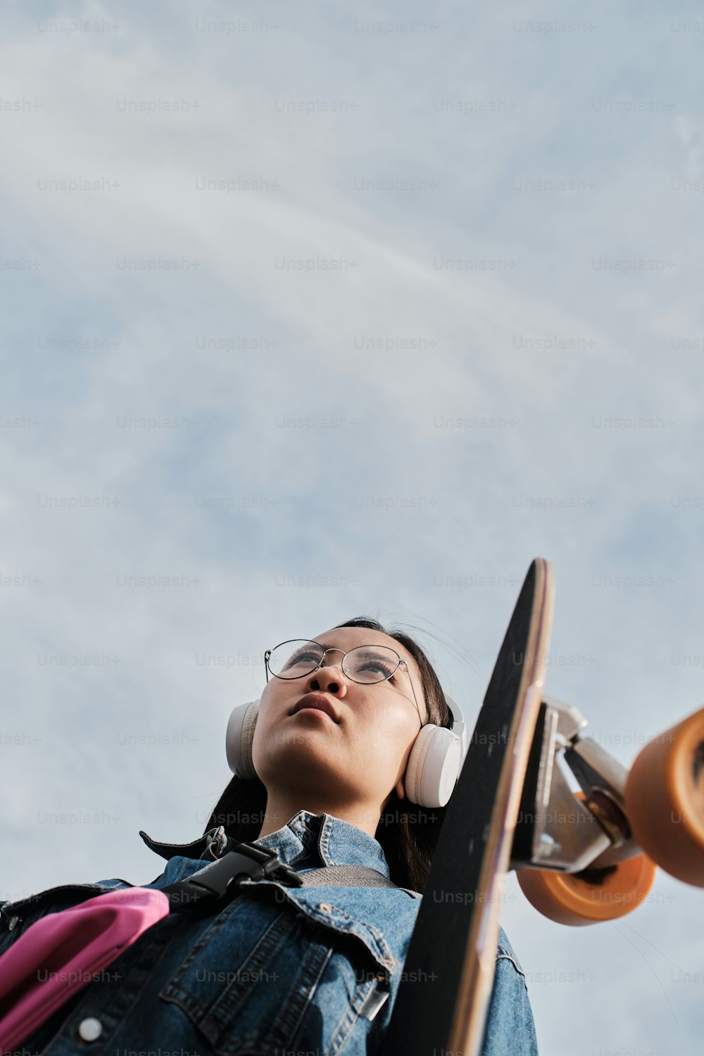 Girl in glasses stands outside with a skateboard shot with sun on the background.