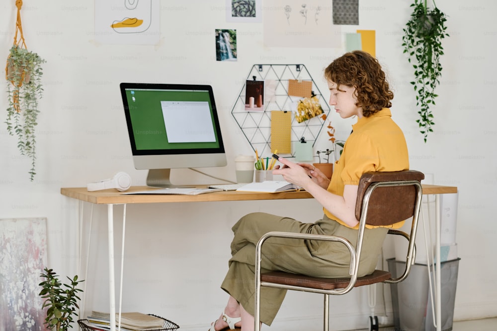 Young serious businesswoman scrolling in mobile phone while sitting in front of computer monitor and organizing work at home