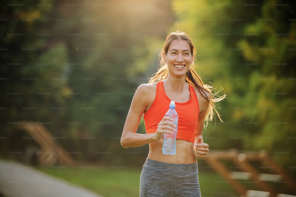 Jovem esportista atraente sorridente positivo com cabelos compridos segurando garrafa de água e correndo em parque público pela manhã.