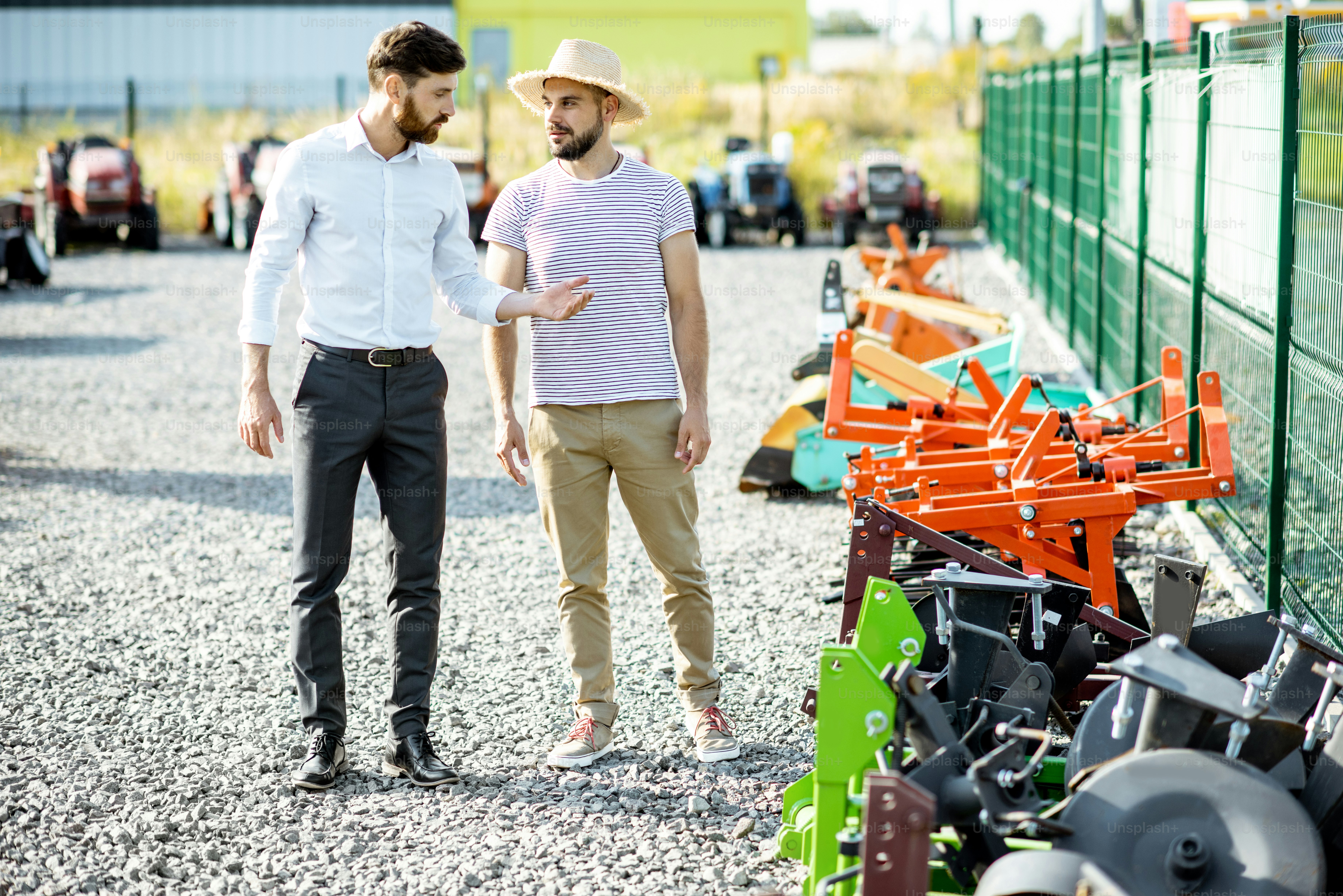 Young agronomist walking with salesman at the open ground of the shop with agricultural machinery, choosing new plow for tillage
