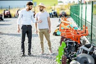 Young agronomist walking with salesman at the open ground of the shop with agricultural machinery, choosing new plow for tillage