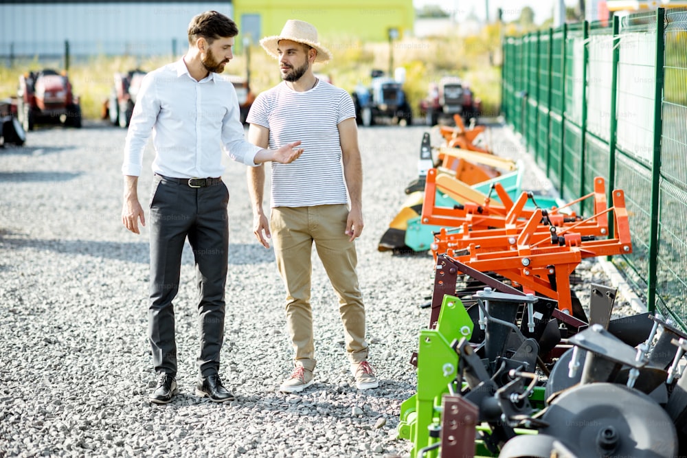 Young agronomist walking with salesman at the open ground of the shop with agricultural machinery, choosing new plow for tillage