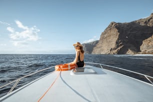 Donna che si gode il viaggio nell'oceano seduta con il salvagente sul naso dello yacht mentre naviga vicino alla costa rocciosa mozzafiato su un tramonto. Concetto di un'attività ricreativa estiva di lusso e di viaggio