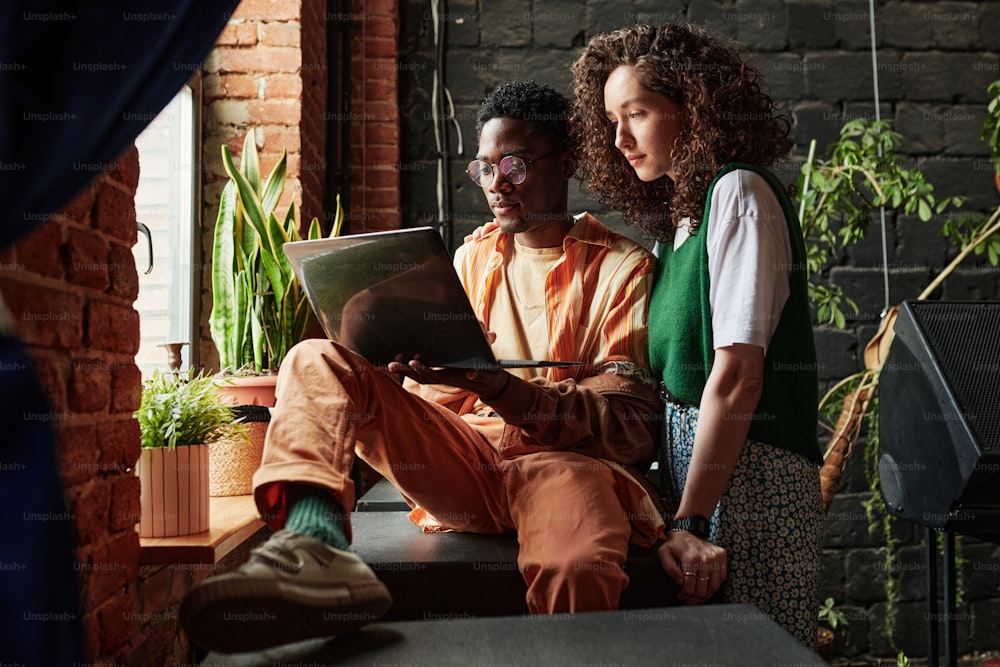 Joven pareja interracial trabajando en la red mientras el hombre sostiene una computadora portátil y su novia de pie junto a él frente a la ventana en el estudio