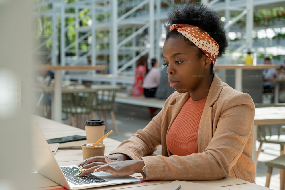 Joven mujer seria de etnia africana en ropa casual inteligente presionando el botón del teclado de la computadora portátil mientras mira la información en línea