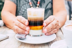 Close up with aged female hands holding a cup of multi coloured coffee for breakfast at the bar - wooden table and bright image - drink and beverage concept for people
