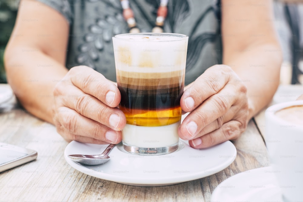 Close up with aged female hands holding a cup of multi coloured coffee for breakfast at the bar - wooden table and bright image - drink and beverage concept for people