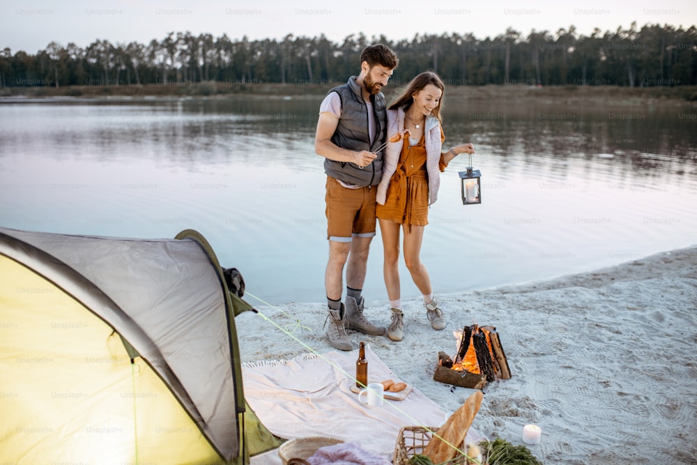 Giovane coppia che si diverte al campeggio sulla spiaggia, in piedi vicino al camino durante la sera