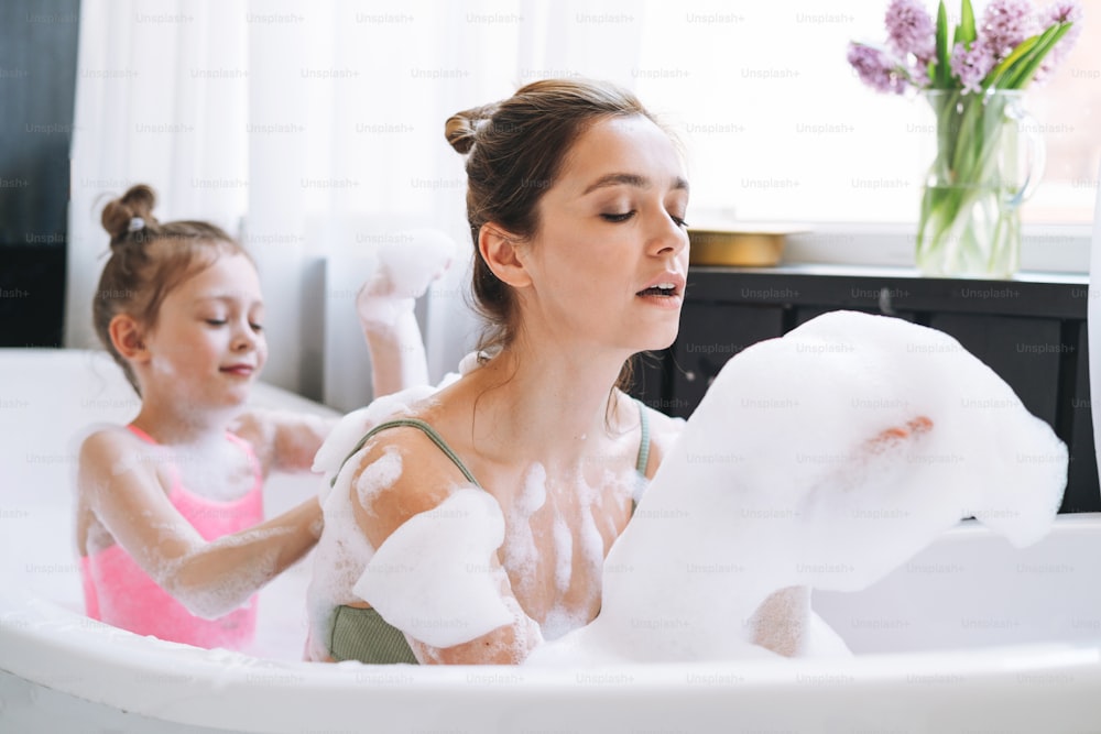Young mother woman and little tween girl daughter having fun in the bath with foam at home