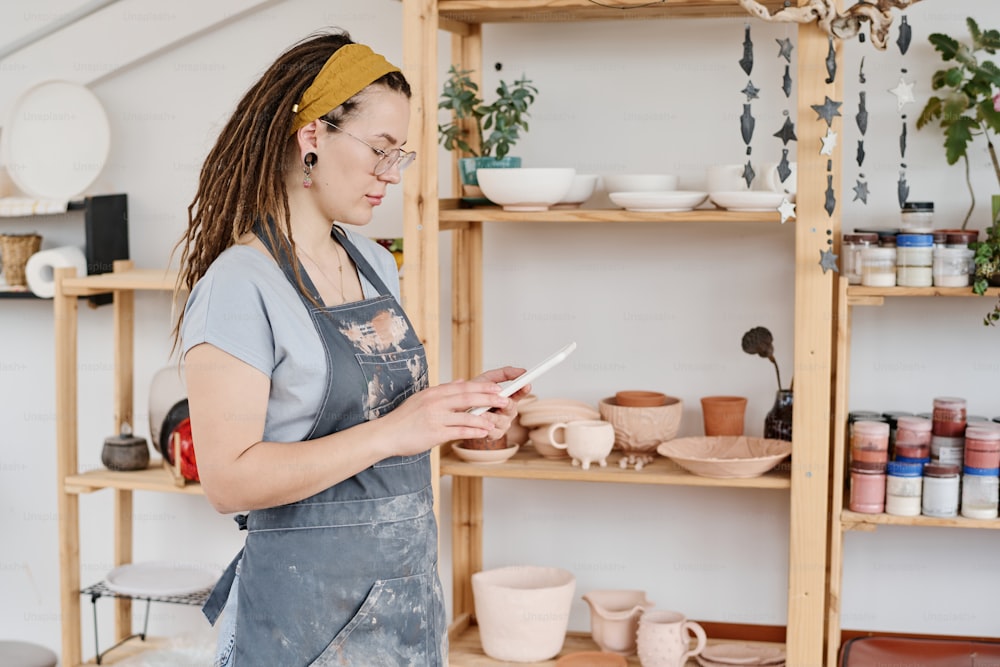 陶器店の若いクリエイティブな女性オーナーがタブレットを使いながら、クライアントの新しいオンライン注文を見ている横から見た写真